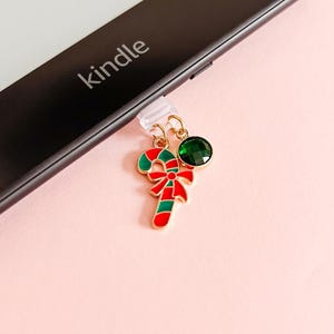 Puede incluir: Un charm dorado con un diseño de bastón de caramelo rojo y verde y una gema verde, adjunto a un dispositivo Kindle negro. El bastón de caramelo tiene un lazo rojo. El Kindle está sobre un fondo rosa.