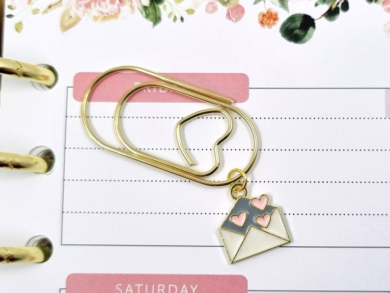 K&ouml;nnte beinhalten: Ein goldfarbener B&uuml;roklammer mit einem kleinen wei&szlig;en Umschlag-Anh&auml;nger mit rosa Herzen. Der Umschlag-Anh&auml;nger ist mit einer goldenen Kette an der B&uuml;roklammer befestigt.