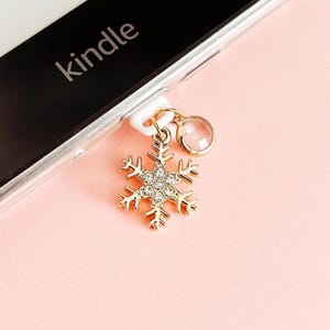 Könnte beinhalten: Ein goldfarbener Schneeflocken-Charm mit klaren Kristallen und einem kleinen, runden, klaren Edelstein hängt an einem weißen Stecker. Der Charm ist an einem schwarzen Gerät mit dem Aufdruck "kindle" befestigt. Der Hintergrund ist ein zartes Rosa.