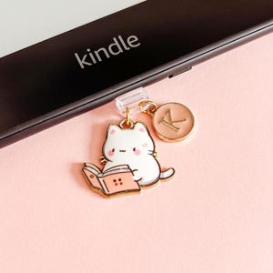 Puede incluir: Un charm dorado con un gato blanco leyendo un libro, un charm con inicial rosa y un clip de fijación transparente. La palabra "kindle" es visible en un dispositivo electrónico negro, sobre un fondo rosa.