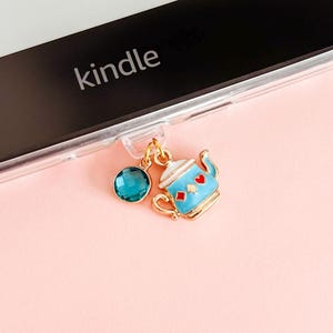 Könnte beinhalten: Ein Kindle-E-Reader mit einem Anhänger. Der Anhänger zeigt eine blaue Teekanne mit roten Herz- und Karo-Akzenten, einen türkisfarbenen Edelstein und goldfarbenes Metall. Das Wort "kindle" ist auf dem E-Reader aufgedruckt.