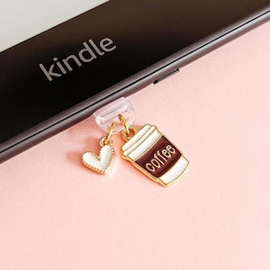 Könnte beinhalten: Ein goldfarbener Anhänger mit einem weißen Herz und einem Kaffeebecher-Design, befestigt an einem Kindle-Gerät. Der Kaffeebecher-Anhänger hat braune und weiße Emaille-Details, mit dem Wort "coffee" darauf gedruckt. Das Kindle-Gerät ist schwarz, mit dem Wort "kindle" darauf gedruckt.
