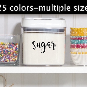 May include: A white plastic container with a clear lid, filled with white sugar. The container has a black label that says "Sugar" in a cursive font. The container is on a white shelf with a glass jar filled with colorful sprinkles and a clear container with colorful cupcake liners.