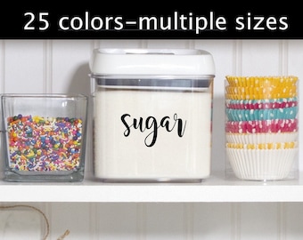 May include: A white plastic container with a clear lid, filled with white sugar. The container has a black label that says "Sugar" in a cursive font. The container is on a white shelf with a glass jar filled with colorful sprinkles and a clear container with colorful cupcake liners.