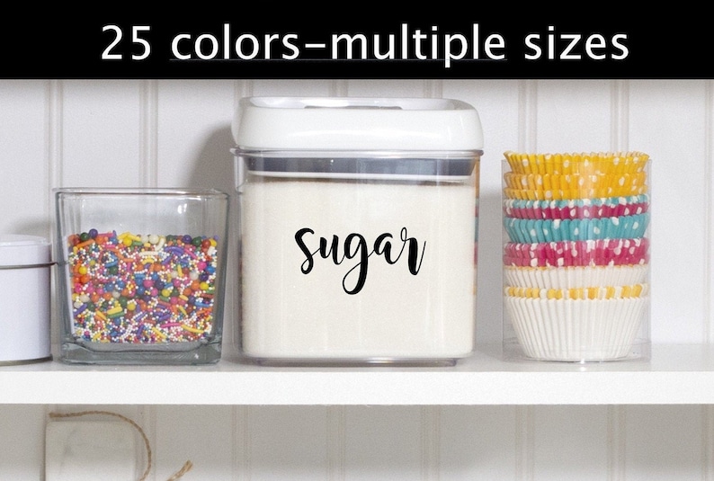 May include: A white plastic container with a clear lid, filled with white sugar. The container has a black label that says "Sugar" in a cursive font. The container is on a white shelf with a glass jar filled with colorful sprinkles and a clear container with colorful cupcake liners.