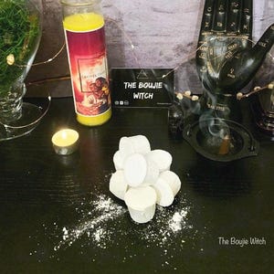 May include: White bath bombs in a pile on a black surface. The bath bombs are round and have a white powder on them. There is a candle, a glass jar, and a black hand with a palm reading on the surface. The text "The Boujie Witch" is on a sign in the image.