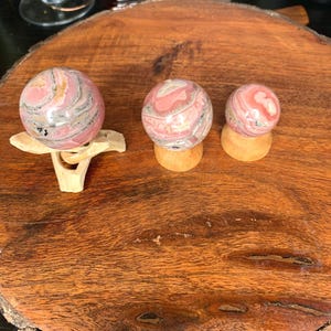 May include: Three pink and gray gemstone spheres of varying sizes, displayed on wooden stands, rest on a large, round, wooden slab. The spheres have a marbled appearance. The wooden slab has a natural, rustic look.