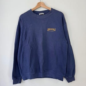 May include: A navy blue crewneck sweatshirt featuring long sleeves and ribbed cuffs. The chest displays the text Aeropostale Sport embroidered in gold lettering. The garment is displayed on a wooden hanger against a plain white background.