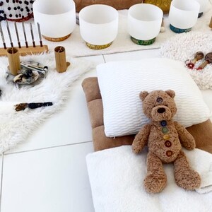 May include: A brown teddy bear with rainbow buttons on its chest is lying on a white pillow and blanket. The bear is on a brown meditation cushion. There are white singing bowls in the background.