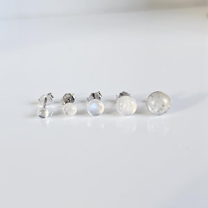 May include: A collection of five pairs of stud earrings with a silver post. The earrings feature round, iridescent, and clear gemstones. The sizes of the gemstones vary from small to large, with the largest having a speckled appearance.