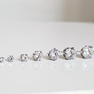 May include: A collection of seven silver stud earrings with clear, round, faceted gemstones. The earrings are arranged in a row, increasing in size from left to right, showcasing the varying gemstone diameters.