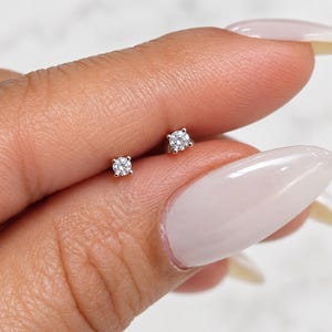 May include: Two small, round, clear stud earrings featuring a single brilliant-cut gemstone set in a four-prong silver-toned metal setting. The earrings are displayed side-by-side, showcasing a minimalist design with a polished finish.