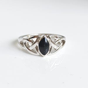 May include: A silver ring with a black, marquise-cut stone at the center. The ring features a Celtic knot design on the band, with intricate interwoven patterns. The ring is set against a white background, highlighting its details.
