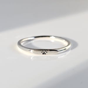 May include: A silver ring with a small paw print design. The ring is thin and has a smooth, polished surface. The paw print is centered on the band.