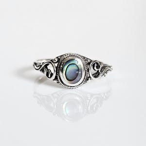 May include: A silver ring with an oval abalone shell center stone. The ring features ornate scrollwork on the band and around the stone. The iridescent shell displays shades of blue, green, and purple. The ring is displayed on a white surface.