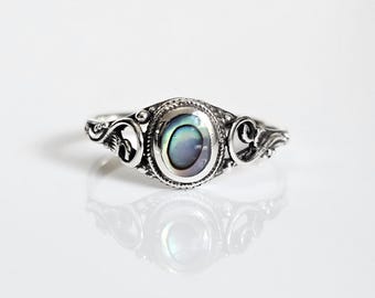 May include: A silver ring with an oval abalone shell center stone. The ring features ornate scrollwork on the band and around the stone. The iridescent shell displays shades of blue, green, and purple. The ring is displayed on a white surface.