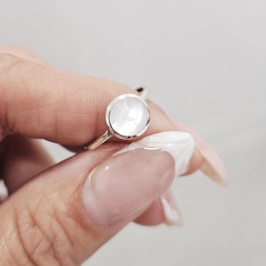 May include: A silver ring with a round, iridescent, white gemstone. The ring's band is thin and polished. The gemstone is set in a simple bezel setting. The ring is shown against a soft, neutral background.