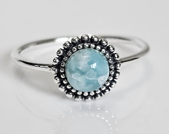 May include: A silver ring with an oval, pale blue gemstone. The gemstone is framed by a beaded edge. The ring's band is slender and polished, reflecting light. The ring is set against a white backdrop.
