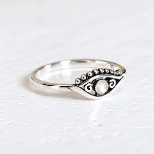 Puede incluir: Un anillo de plata con una pieza central en forma de ojo y una piedra redonda blanca en el centro. El diseño incluye pequeños detalles de cuentas en el borde superior y adornos en espiral a los lados del motivo, hecho de metal pulido.