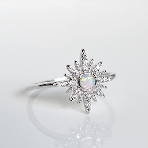 Puede incluir: Un anillo de plata con un diseño de estrella. El anillo presenta una piedra central redonda de ópalo iridiscente, rodeada de piedras preciosas claras más pequeñas. El diseño de estrella irradia hacia afuera, creando un efecto radiante. El anillo está sobre un fondo blanco.