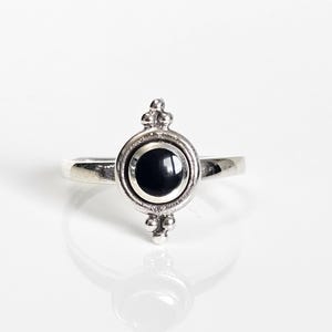 May include: A silver ring with a round, black stone set in a decorative bezel. The ring features a simple band and ornate detailing around the stone, with small, raised accents. The ring is displayed on a white surface, reflecting the light.