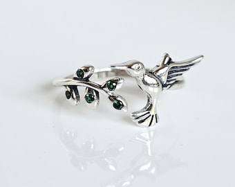 Könnte beinhalten: Silberring mit einem Kolibri, der auf einem Blattzweig mit grünen Edelsteinen sitzt.