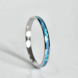May include: A silver ring with a band of inlaid blue opal. The opal has a mix of light and dark blue hues. The ring is set against a white background.