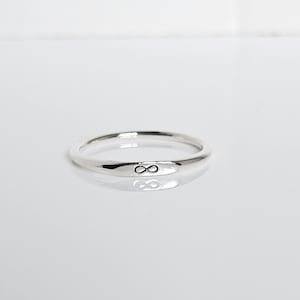 May include: A silver ring with an infinity symbol engraved on the band. The ring is smooth and shiny, reflecting the light. The infinity symbol is a small, dark marking on the front of the ring.
