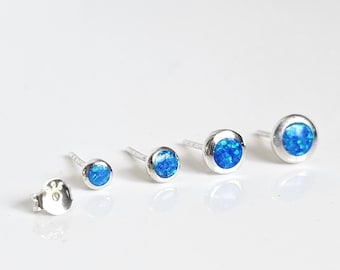 May include: A collection of five silver stud earrings with round, bright blue opal stones. The earrings vary in size, with the smallest on the left and the largest on the right. The earrings are displayed on a white surface.