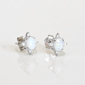 May include: A pair of silver stud earrings. Each earring features a central oval-shaped white stone surrounded by smaller clear stones. The earrings have a starburst design and are set against a white background.