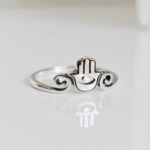 May include: A silver ring featuring a Hamsa hand design. The ring has a detailed open hand with a central eye symbol, flanked by decorative swirls. The ring is displayed on a white surface, reflecting the ring's design.