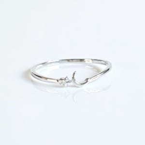 May include: A thin silver-toned ring featuring a small crescent moon and a tiny star-shaped accent. The star is set with a clear, sparkling stone. The band is smooth and polished, creating a delicate and reflective metallic appearance throughout.