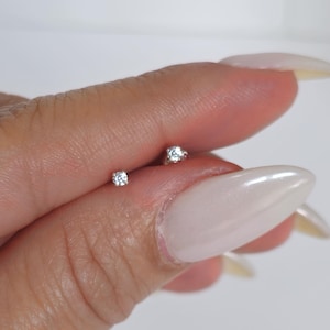 Puede incluir: Un par de pequeños pendientes de botón redondos, cada uno con una sola gema transparente. Los pendientes se sostienen entre los dedos sobre un fondo blanco. Las gemas brillan, reflejando la luz.