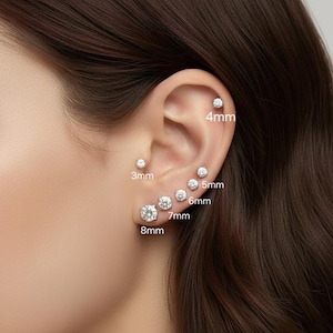 May include: A series of clear, round stud earrings of varying sizes, from 3mm to 8mm, are displayed on an ear. The earrings are set in a light-colored metal and are arranged in a descending size order, with the largest at the bottom.