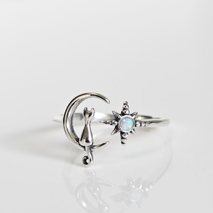 May include: A silver ring featuring a crescent moon with a cat silhouette and a starburst design. The starburst has a central opal-like stone. The ring is set against a white background.