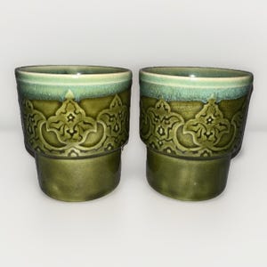 May include: Two ceramic cups with a green and teal glaze. The cups have a decorative embossed pattern around the middle and a solid green base. The rims are teal, and the cups are set against a white background.