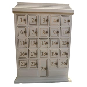 May include: A white wooden advent calendar with 25 numbered drawers. The numbers are in a gold color and are displayed in a grid pattern. The calendar has a small roof and a base.