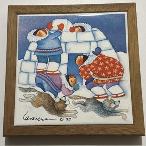 Op de afbeelding: Een ingelijste keramische tegel met een Inuit dorpsscène. Het kunstwerk toont figuren die een iglo bouwen, met rennende honden op de voorgrond. De tegel is geplaatst in een houten frame. Gesigneerd "Canuck" en gedateerd "95".