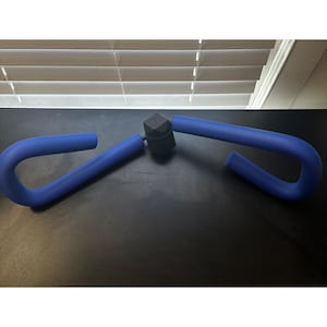 Pode incluir: Um exercitador de coxas azul com um conector central preto. As alças curvas são de um azul vibrante, projetadas para exercícios de pernas. O dispositivo é mostrado em uma superfície escura.