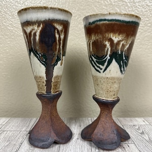 Puede incluir: Dos copas de cerámica hechas a mano. Las copas tienen una parte superior acampanada con un esmalte blanco, marrón y verde. La base es un diseño marrón oscuro de tres patas. Las copas se exhiben sobre una superficie de madera.