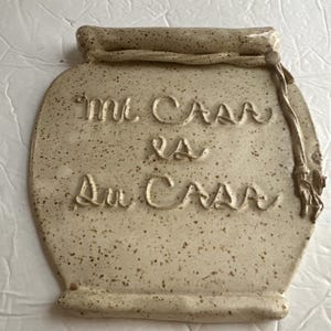 May include: A speckled, off-white ceramic plaque in the shape of a jar, with the words "Mi Casa es Su Casa" embossed on the front. The plaque has a rolled top and a decorative rope detail on the side.