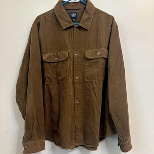 May include: A brown long-sleeve corduroy shirt featuring a standard collar, a front button-down closure, and two chest pockets with buttoned flaps. The shirt includes a GAP brand label at the inner neckline and matching brown buttons throughout.