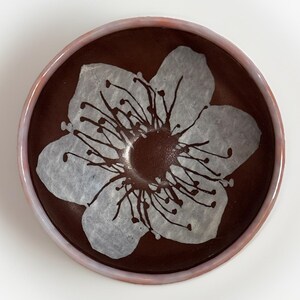 May include: A round, ceramic bowl with a dark brown interior and a light pink rim. The bowl features a large, white floral design in the center with dark brown accents. The design is reminiscent of a cherry blossom.