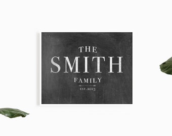 May include: A framed chalkboard-style sign with the text "THE SMITH FAMILY EST. 2013" in white lettering. The background is a dark gray, textured surface. The sign is centered on a white background, with green leaves in the corners.