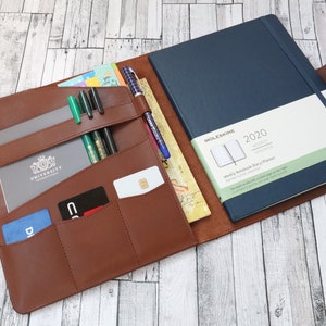 Può includere: Una cartella porta-agenda in pelle marrone con più tasche e fessure per penne, carte e un taccuino. La cartella è aperta per rivelare un'agenda settimanale Moleskine 2020 blu.