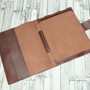 May include: A brown leather folder with a flap closure and a pen holder. The folder is open and the inside is lined with a light brown fabric.