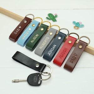 Puede incluir: Una colección de llaveros de cuero personalizados en varios colores, incluyendo marrón, azul, verde, gris, marino y rojo. Cada llavero presenta un anillo de metal plateado y está grabado con un nombre o palabra. También hay un llavero de cuero negro con una llave de coche.