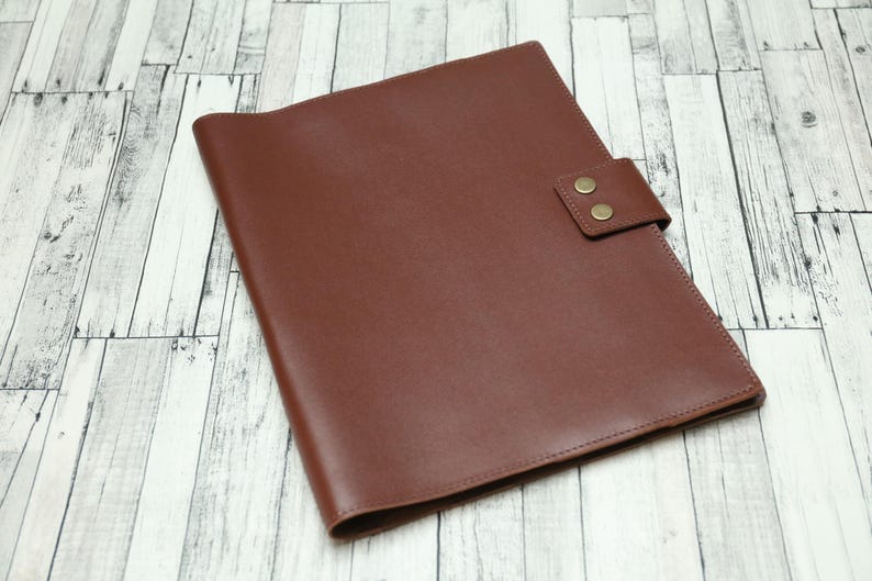 May include: A brown leather folder with a flap closure and two brass button accents.