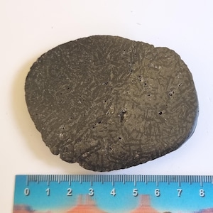May include: A dark gray, irregularly shaped stone with a textured surface and a pattern of intersecting lines. The stone is placed on a white surface next to a ruler showing measurements in inches.