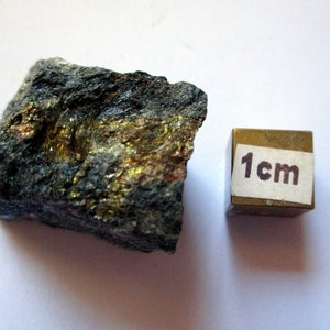 May include: A rough, dark gray rock with patches of shimmering gold and bronze. A small, brass cube with a 1 cm label is next to the rock, providing scale. The rock's surface is textured and uneven.